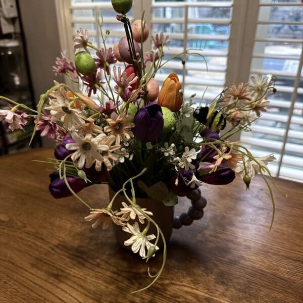 A colorful flower arrangement in a vase on a wooden table near window blinds.